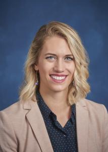 Headshot of Adrienne Woods with long blonde hair in front of a blue background wearing a tan suit jacket.