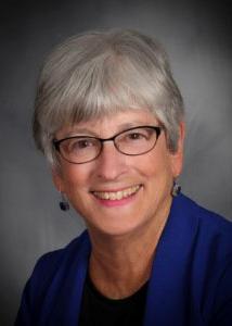 Headshot of Carol Weisman in front of a grey background wearing glasses, statement earrings and a blue jacket.