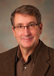 Headshot of David Almeida in a brown dress shirt and jacket wearing glasses.