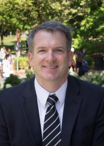 Headshot of David Conroy with grey, short hair wearing a suit and striped tie (picture taken outdoors).