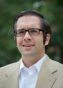Headshot of Douglas Wrenn with short dark hair outside wearing tan suit jacket and glasses.