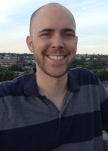 Headshot of Jonathon Daw (outdoors) wearing a black and grey collared shirt.