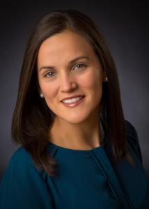 Headshot of Susana Quiros in front of a grey backdrop with long, brown hair wearing a dark blue top.